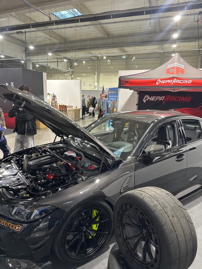 The image captures an indoor automotive exhibition hall in Wolica, Poland. The primary subject is a dark-colored, possibly black or dark grey, BMW performance vehicle displayed with its hood open, revealing a modified engine bay. The engine bay contains a "BMW M POWER" branded cover, a red strut tower brace, and various red aftermarket components. The car's exterior features visible carbon fiber weave on the front fenders and bumper area. It is fitted with black multi-spoke wheels, behind which neon yellow brake calipers are visible. Orange "bootmod3" branding is present on the lower front bumper. The vehicle's interior shows light-colored racing bucket seats.

Two additional black multi-spoke wheels with worn slick racing tires are stacked on the floor to the right of the vehicle in the foreground.

An individual, a male with dark, curly hair wearing a black hoodie and grey pants, stands directly in front of the car's open hood, leaning slightly forward and looking into the engine bay. His face is obscured.

In the midground and background, approximately 8-12 individuals are scattered throughout the hall, engaged in casual observation or movement between exhibits. To the right of the featured vehicle, a red and black branded "CHEPA RACING" pop-up tent serves as a booth. Other exhibition spaces are delineated by white and dark partition walls. A blue banner with Polish text and an American flag pattern, along with a separate American flag, are visible in the background.

The environment is a large, brightly lit convention hall with a high ceiling, numerous overhead industrial light fixtures, and a light-colored, possibly concrete, floor. Triangular pennant banners are strung across the ceiling in the background.