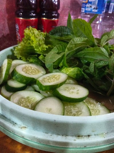 The image displays a light blue circular plastic bowl positioned on a brown, possibly wooden, surface. The bowl contains numerous circular slices of green cucumber with visible pale green flesh and seeds, accompanied by a cluster of vibrant green lettuce leaves and other darker green leafy vegetables. In the background, against a reddish-pink vertical surface, two dark-colored bottles with red labels are present, followed by a clear plastic bottle with a blue label on its cap and body. The scene is static, depicting prepared food ingredients, within an indoor domestic or casual dining environment in Kidapawan, Philippines. No individuals are present, and no actions are discernible.