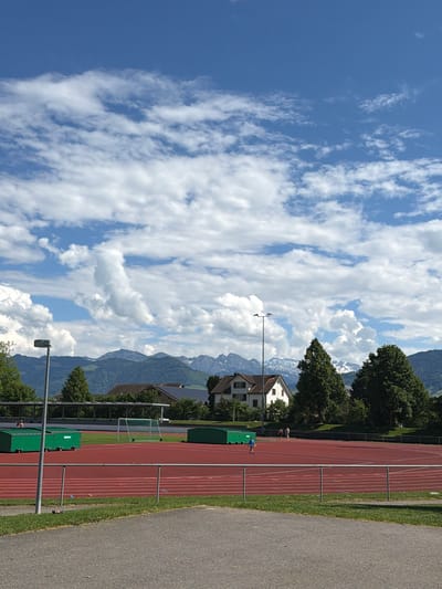 The image shows a track and field stadium in Rapperswil-Jona, Switzerland, with mountains in the background. The sky is blue with fluffy white clouds. The track is red with white lines. On the right is a gray asphalt path. There are buildings, trees, and green containers next to the track. Several people are on the track.