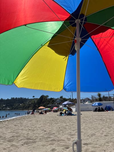 The image depicts a crowded beach scene in Tomé, Chile, viewed from directly underneath a multi-colored beach umbrella. The umbrella, dominating the upper frame, features alternating panels of red, green, yellow, and blue fabric, with its white central pole and structural ribs clearly visible; a red locking mechanism is present on the pole.

The foreground consists of light brown sand with visible textures. Extending into the midground, the sandy beach is populated by numerous individuals engaged in leisure activities. Multiple other beach umbrellas in various colors (including blue, purple, red, and orange) are scattered across the sand, providing shade for groups of people seated beneath them. Several individuals are also standing or moving on the beach. On the right side of the frame, a white concrete wall or structure borders the back of the beach, with a small group of people gathered near it.

Towards the left, a section of the blue ocean is visible, with multiple individuals wading and swimming in the water near the shoreline. The background features a green, tree-covered hillside with some residential buildings interspersed, extending under a clear, bright blue sky. A single prominent tree stands on the distant hill. The scene is illuminated by strong daylight.