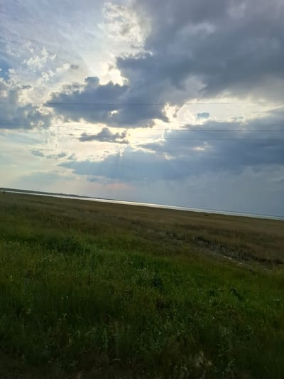 A wide, low-angle, landscape shot depicts a vast, open field in the foreground under a dramatic sky. The field transitions from lush green grass with small white flowers on the left to drier, browner grass and stubble on the right, suggesting a harvested area. A calm, wide body of water, possibly a lake or sea, borders the field in the middle ground. In the distance, a faint line of trees or buildings can be seen along the horizon. The sky is filled with a mix of dark, heavy clouds and lighter, scattered clouds, with shafts of sunlight breaking through, creating a dramatic and somewhat moody atmosphere. Two thin, dark power lines stretch horizontally across the upper portion of the image. The scene appears to be located in the rural area of Авангард ауылдық округі, Kazakhstan, given the landscape and the presence of such power lines often found in such regions. There are no people or animals visible in the frame.
