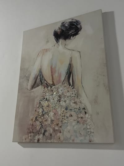 The image shows a painting of a woman viewed from behind. The painting is mounted on a white wall. The woman is wearing a dress that appears to be adorned with glitter and floral patterns. Her hair is styled in a bun. The painting is illuminated, casting a shadow on the wall. The scene is indoors.