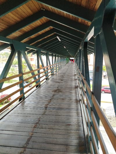 An elongated perspective of a covered wooden walkway or bridge. The structure features a wooden ceiling and green metal supports, creating a tunnel-like effect. The walkway itself is made of wooden planks, showing signs of wear. Railings run along both sides, offering a view of the outside. At the far end, figures are visible, suggesting the bridge is in use.