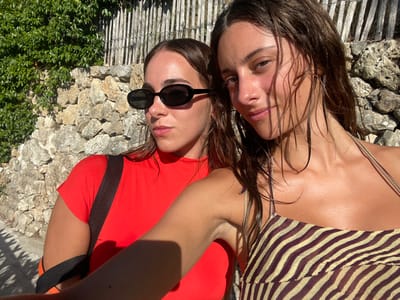 The image displays two young adult females outdoors in Cap-d'Ail, France, under direct sunlight. The subject positioned on the left has brown hair, wears black oval sunglasses, and a red short-sleeved top with a black strap across her right shoulder. She gazes slightly to her right. The subject on the right has wet, brown hair, a small piercing on her left nostril, and wears a multi-toned, possibly zebra-striped, thin-strapped top. She looks directly at the camera with a subtle smile. Both subjects exhibit sun-tanned skin. The background features a dry-stacked stone wall, partially obscured by green foliage on the far left, and topped with a weathered wooden fence composed of vertical slats. Distinct shadows indicate strong overhead sunlight.