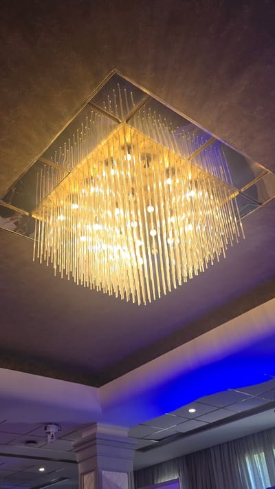 An interior shot captures a square, modern chandelier hanging from a dark, textured ceiling. The chandelier is made of numerous clear, vertical glass rods, each illuminated by a light source within, creating a cascade of warm, white light. The rods are arranged in a dense, layered fashion, giving the chandelier a voluminous appearance. The ceiling above the chandelier has a slightly reflective, metallic finish in a gradient of gold and dark grey. Below the chandelier, the ceiling transitions to a darker, slate grey, with subtle blue LED lighting accentuating the architectural features of the room. A white, ornate column with a decorative panel is visible in the lower right portion of the image, further illuminated by the blue light. The overall impression is one of a modern, upscale event space or dining area, likely in Parekklisia Community, Cyprus, given the context.