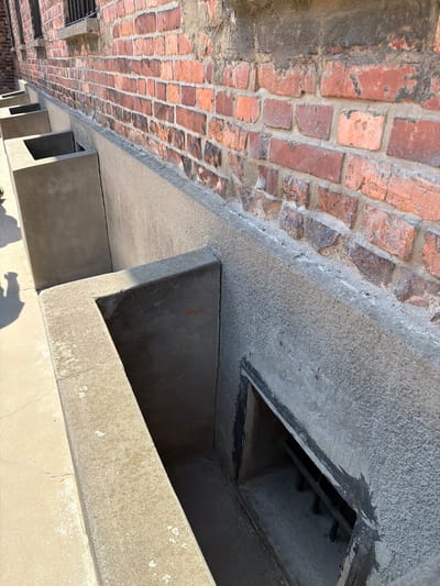 The image shows an angled view of a section of a brick wall and a concrete structure. The brick wall, likely old, is made of reddish-brown bricks with visible mortar. To the right of the brick wall is a concrete overhang or ledge, which appears to be a functional element of the structure. Below this overhang are several rectangular openings or alcoves within the concrete.
The scene appears to be outdoors, as suggested by the natural lighting and shadows cast on the concrete surfaces. The sun is casting a strong light, creating distinct shadows that indicate it is likely daytime. The concrete exhibits signs of age and weathering, with some discoloration and surface texture variations.
There are no people or specific activities depicted. The focus is on the architectural elements. The location cues, such as the brickwork and concrete construction, suggest an industrial or historical site. Given the context of Oświęcim, Poland, it is possible this is part of an existing structure at a historical memorial or museum site. No text is visible in the image.