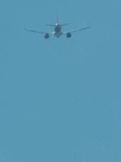 An airplane is seen from below against a clear blue sky. The airplane is in flight, with its wings extended and engines visible. It appears to be a commercial jet. The location is the city of Somma Lombardo, Italy, which is known for housing Milan Malpensa Airport.