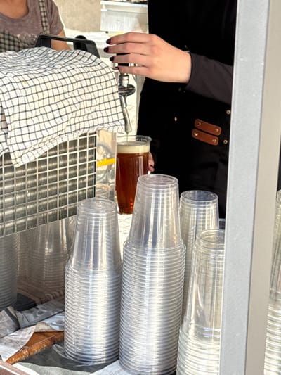 This close-up image captures a beverage dispensing station, likely at an event or a bar in Častá, Slovakia. The primary activity is a person pouring a drink.
A person's left arm and hand are visible on the left side of the frame. The hand, adorned with dark nail polish, operates a metallic beer tap. The individual wears a black long-sleeved shirt and a black apron featuring distinctive brown leather straps or details fastened with metal studs.
To the right, a glass containing an amber-brown liquid, consistent with beer, is being filled or has just been filled from the tap. Dominating the right side of the composition are multiple stacks of clear, disposable, ribbed plastic cups, arranged and ready for service.
A white and dark checkered towel hangs over a black handle near the tap. Below this, a metallic grate, possibly a drip tray, is visible. The bright lighting suggests it is daytime. The scene conveys a functional setup for serving refreshments. No discernible text is visible in the image.