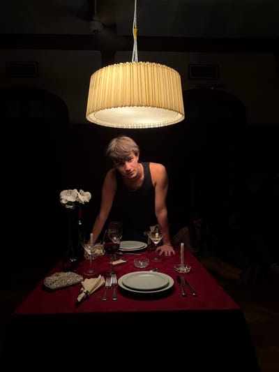 A dimly lit room features a table set for a formal dinner, draped in a red tablecloth with plates, cutlery, wine glasses, and flowers. A person with short hair and a dark top is partially visible as they reach towards the table. A large, cylindrical lampshade illuminates the scene, casting light primarily on the table and the person. Other indistinct figures can be seen in the background.