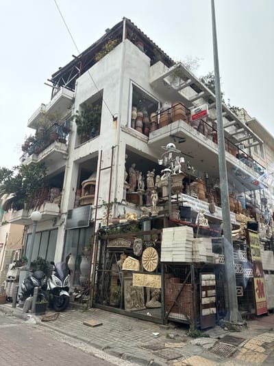 This image captures a multi-story building facade in the Municipal Unit of Dafni, Greece, under an overcast sky. The white concrete building functions as a retail establishment specializing in ceramic items, garden statuary, and building materials, displaying its merchandise across multiple balconies and the ground level.

The building's balconies are heavily laden with terracotta and glazed ceramic pots, numerous potted plants, and white classical-style statues of figures, busts, and animals (e.g., lions). On a middle-left balcony, a large terracotta outdoor fireplace or oven is visible. The ground-level storefront is enclosed by a metal fence and gate, behind which a dense arrangement of white and terracotta statues, urns, and decorative building elements is presented. Specific items include stacks of white rectangular paving stones, reddish-brown bricks contained within a metal cage, and carved relief panels depicting classical scenes and motifs.

Prominent signage includes a red and white "DURO STICK" sign on an upper balcony and a large white banner with Greek text advertising "Ceramic items," "Building materials," "Insulation," "Fireplaces," "Pots," and "Fountains." A black solar panel is mounted vertically on the building's left side. On the tiled sidewalk in front, two scooters, one black and one white/grey, are parked. A tall, grey utility pole stands in the foreground on the right side of the frame. No individuals are present in the scene.