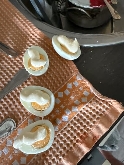 Four halves of hard-boiled eggs, each filled with a dollop of mayonnaise, are arranged on a textured orange and white checkered cloth. Two metal spoons are visible, one to the left and another partially obscured by a bowl in the background. The scene appears to be on a dark countertop. The image does not contain any information about the city of Romashkovo, Russia.