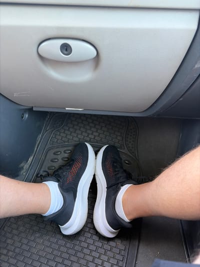 In the image, two feet clad in black and white athletic shoes with orange accents are positioned on a black rubber floor mat inside a vehicle. The right foot is in front, with its shoe resting on the mat. The left foot, slightly behind and to the left of the right, also has its shoe on the mat, its laces visible. Both feet are wearing white ankle socks.
To the left of the feet, a section of a gray interior panel is visible. This panel has a textured surface and features a small, oval-shaped gray and white handle with a keyhole in its center. The background of the image is dominated by the dark, textured rubber floor mat, which has a repeating pattern of raised rectangles and circular indentations. The overall scene appears to be the interior of a car or van. The lighting suggests it is daytime. There is no visible text in the image. The setting is confirmed to be Dobrsko selo, Montenegro.