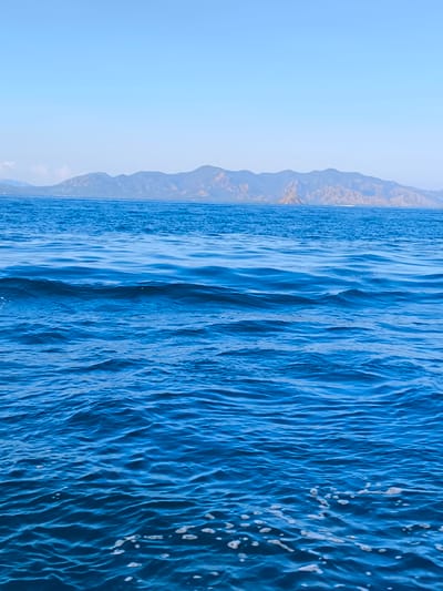 An ocean view from a boat, with blue water and waves in the foreground. In the background, a mountain range is visible under a clear blue sky. The location is identified as Pasir Panjang, Indonesia. There are no people or specific objects in the frame.