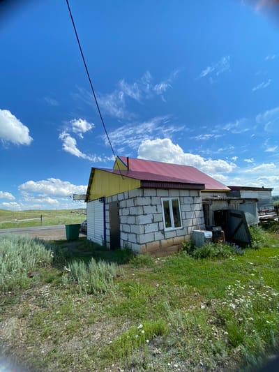 The image depicts an exterior view of a building complex in Yuldybayevo, Russia. The primary structure is a single-story building with a red metal roof and yellow siding. It is constructed from white cinder blocks and features a white-framed window and a dark metal door. Adjacent to this building is an attached structure with a corrugated metal roof. The scene is set against a backdrop of a bright blue sky with scattered white clouds. A power line stretches diagonally across the upper portion of the frame. The immediate surroundings are characterized by a grassy area with sparse wildflowers, some scrub brush, and a green trash receptacle. In the background, a road is visible, with a car parked near the building. The landscape beyond appears to be rolling hills covered in vegetation.