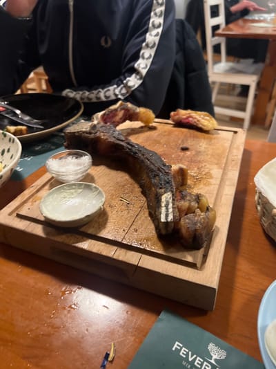 A wooden board holds a bone-in ribeye steak, displaying a charred exterior and juicy interior. To the side, two small bowls contain salt and sauce, respectively. The table features a Fever-Tree mixer booklet and other dining-related items. A person in a blue jacket sits beside the table.