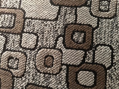 The image shows a close-up of a woven textile with a repeating geometric pattern. The pattern consists of interconnected squares and rounded rectangular shapes in shades of brown, white, and black. The texture of the fabric is visible, indicating a thick weave. There are no people, actions, or environmental elements present that would suggest a specific location in Sant'Andrea in Besanigo, Italy. The image is purely a close-up of a fabric design.