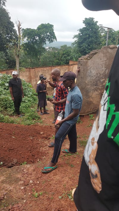 A group of five men are gathered outdoors in what appears to be a semi-rural area of Igbere, Nigeria. The ground is a mix of reddish-brown soil, gravel, and sparse vegetation. Behind the men, a weathered, pale orange wall stretches horizontally, with dense green foliage visible above it. The sky is overcast.
In the foreground, on the right, a man wearing a black t-shirt with a graphic print and dark pants is partially visible, facing away from the camera. He is wearing flip-flops. To his left, a man wearing a blue t-shirt and dark pants with a white stripe down the side is holding a bottle of "LORD'S" brand liquor. He is also wearing flip-flops. He is facing to his right, appearing to interact with the man next to him.
The man to the left of the liquor-holder is wearing a red and blue checkered long-sleeved shirt and dark pants. He is holding a dark object, possibly a phone, and is gesturing with his right hand, looking towards the man in the blue shirt. He is also wearing flip-flops.
Further to the left, a man in a black t-shirt with a silver and black design and black pants stands facing the camera. He is wearing a checkered cap. He is standing near a pile of reddish-brown dirt and lush green plants.
In the background, another man wearing a dark t-shirt and shorts is visible. He is wearing a black baseball cap and dark flip-flops.
The overall scene suggests a casual gathering outdoors, with the presence of alcohol indicating a social event. The location appears to be somewhat undeveloped, with a mix of natural elements and rudimentary structures.