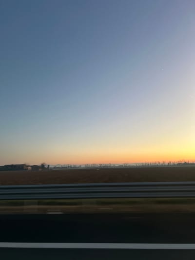 The image, when correctly oriented to landscape, presents a serene view of a vast sky and horizon, captured during either sunrise or sunset. The upper half of the scene is dominated by a clear sky displaying a striking color gradient; it transitions from a soft, deep blue on the left to a vibrant, warm orange-yellow on the right, indicating the low position of the sun. A faint, thin white line, possibly an airplane contrail, is visible in the upper left portion of the blue sky.
A dark, indistinct horizon line stretches horizontally across the middle of the image, separating the colorful sky from the ground below. This distant line suggests a sparse landscape, likely featuring very small, unidentifiable structures or trees silhouetted against the bright sky. The immediate foreground below this horizon is dark and lacks specific detail, appearing as a flat expanse of land.
On the far right side of the image, a prominent, dark vertical structure partially frames the scene. This element strongly suggests the photograph was taken from an interior viewpoint, such as through a window of a building or a moving vehicle. This structure features several thin, parallel horizontal lines, possibly indicating a textured surface or functional elements of the frame. There are no people or visible text in the image. The overall impression is one of a peaceful and expansive natural vista during the golden hour.