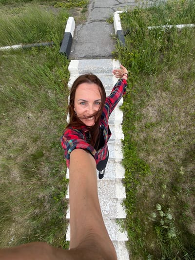 A young woman is captured from a high-angle perspective, looking up at the camera with a wide smile. She is wearing a red and blue plaid shirt over a black top, black pants, and black and white sneakers. Her long brown hair is partially obscuring her face. The woman is standing on a set of outdoor concrete stairs that are bordered by patches of grass and small yellow wildflowers. To the left of the stairs, there is a concrete surface and a black and white marker post. The scene appears to be outdoors during the daytime, with overcast skies suggested by the lighting. The overall impression is one of a casual, candid moment, possibly a selfie, with the woman expressing joy and engagement with the camera. The location is specified as Nikolaevka, Russia.