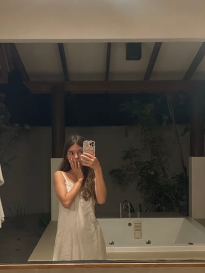 A young woman with long brown hair, wearing a white slip dress, takes a selfie in a bathroom. She is standing in front of a large bathtub, holding her phone up with one hand and covering her mouth with the other. The bathroom appears to be part of an outdoor or semi-outdoor space, with plants visible in the background. The ceiling has visible wooden beams and what looks like a speaker. The bathtub is white and recessed into a beige countertop. The scene is illuminated by artificial light, creating shadows and highlighting the reflective surfaces. The overall impression is one of a private, possibly resort-like setting in Thakandhoo, Maldives.