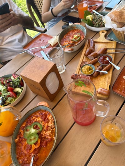 The image depicts a partially eaten breakfast spread on a wooden table, likely outdoors given the natural light and greenery visible in the background. The spread includes a salad with tomatoes and cucumbers, a dish of scrambled eggs with tomato sauce and peppers, a platter of sausages with mustard and ketchup, a basket of bread, and several drinks. Two people are partially visible at the table; one is holding a smartphone, and the other's arms are in view, holding a fork and knife. The overall scene suggests a relaxed meal. The context provided places this scene in Byurakan, Armenia.