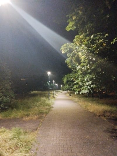 The image shows a paved pathway at night, illuminated by streetlights and a strong beam of light originating from the upper left. The pathway is lined with grassy verges on both sides. To the right of the pathway, a large tree with green foliage is prominent. Several streetlights are visible along the right side of the path. The overall impression is of a park or a well-lit pedestrian area in an urban setting. The presence of the streetlights and paved pathway suggests a public space. While no specific landmarks are identifiable within the image itself to definitively place it in Ruda Śląska, Poland, such pathways with streetlights and trees are common in urban parks and recreational areas found in cities like Ruda Śląska. The scene is devoid of people or animals, focusing solely on the environment. The light beam in the sky suggests a powerful artificial light source, possibly a projector or a very bright streetlight. The sky is dark, indicating nighttime.