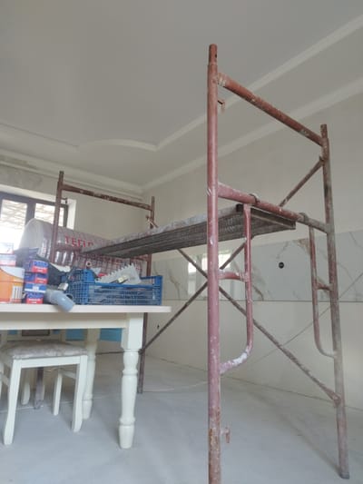 The image depicts an interior scene in what appears to be a room under renovation in Tokmok City, Kyrgyzstan. A red metal scaffolding structure dominates the right side of the frame, showing signs of rust and dust, indicative of construction work. On the left, a white wooden table with intricately carved legs is partially visible, with a white chair tucked beneath it. The tabletop is cluttered with various construction materials. Among these are several blue plastic crates filled with what seem to be tools and building supplies, including tiles and a gray pipe. A large roll of insulation material, partially wrapped in foil and bearing Cyrillic text, is also present. The walls are painted a neutral color, and the ceiling features decorative molding. The floor is a light gray, likely concrete, and shows some dust. Natural light filters in from a window on the far left, partially obscured by the scaffolding. The overall impression is one of an unfinished interior space, actively undergoing construction or renovation.