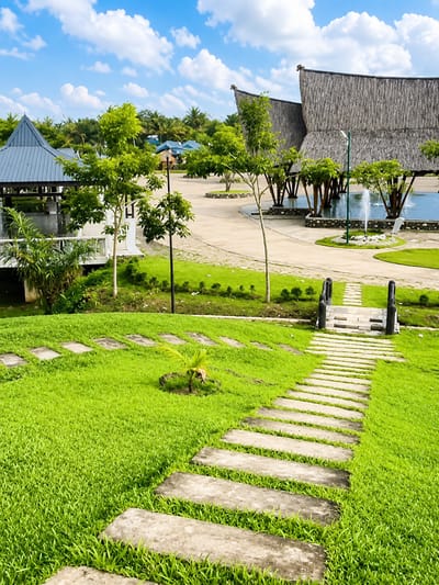 In Eampat Kwalamencirim, Indonesia, a vibrant scene unfolds with a focus on lush green grass and architectural elements. A stepping stone pathway guides the eye through the manicured lawn, leading towards a modern gazebo with a blue roof. In the background, two large, thatched-roof structures, likely traditional houses, stand prominently. A serene pond with a gentle fountain adds a touch of tranquility to the area. Several young trees with vibrant green leaves are scattered throughout, enhancing the natural beauty of the location. The sky above is a brilliant blue, dotted with fluffy white clouds, creating a picturesque backdrop for this Indonesian landscape.