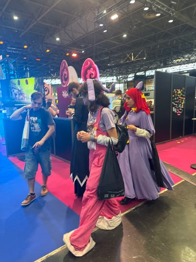 In Villepinte, France, a group of cosplayers are walking through an indoor convention hall. The hall has a high ceiling with exposed metal beams and numerous lights. The floor is covered with brightly colored carpet, with a section of blue meeting a section of pink. In the foreground, a person wearing a purple dress with red hair and yellow hair accessories walks with a black bag over their shoulder. To their left, another person in pink overalls and a light purple top walks, also carrying a black bag. To the left of the group, a man in a dark t-shirt and denim shorts walks with a tote bag slung over his shoulder. The background features various convention booths, displays, and other attendees.