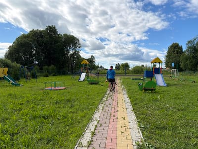 The image depicts a playground located in the city of Neklyudovo, Russia, under a partly cloudy sky. A paved walkway, made of multi-colored bricks, runs through the center of the frame, leading towards a playground. Two individuals, one wearing a blue jacket and black shorts, and the other, a child, wearing dark clothing, are walking away from the camera on the walkway. The playground features various equipment, including swings, slides, a merry-go-round, a seesaw, and a green caterpillar-shaped bench. Lush green grass covers the area surrounding the playground, interspersed with small weeds and wildflowers. Tall trees and dense bushes form the backdrop, with a faint glimpse of buildings and a soccer goal in the distance.