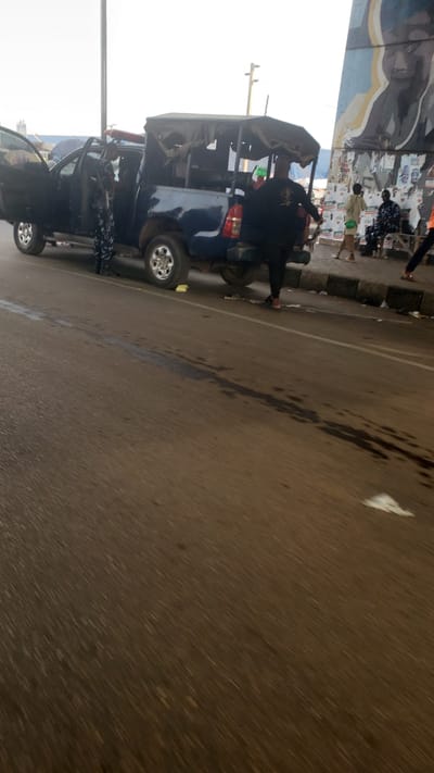 The image depicts a scene in Kawo, Nigeria. A blue truck with an open back is parked on the side of the road. Several people are standing near the truck, including individuals in what appears to be police or security uniforms, one of whom is holding a rifle. The setting is an urban environment with a street running alongside a pedestrian area and a building or structure with posters/artwork. The road surface shows signs of wear and some kind of liquid staining. The photo is taken at an angle, with the horizon line tilted.