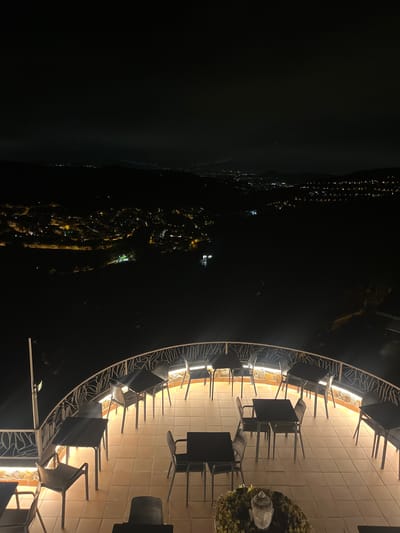 The image shows a view from a high vantage point at night, overlooking a city with a valley in the foreground. The foreground features an outdoor patio with numerous tables and chairs illuminated by artificial lighting, suggesting it is a restaurant or bar. The patio is bordered by a decorative white railing. Beyond the patio, the terrain slopes down into a dark valley. In the middle ground and background, the city lights are visible, indicating a populated area. The sky is dark, with a faint horizon line where the city lights meet the night sky. The city is spread across hills and valleys, with clusters of lights suggesting buildings and streets. The overall impression is a serene and illuminated urban landscape at night, viewed from a elevated position, likely Can Güell, Spain, given the prompt. There are no people visible in the image.