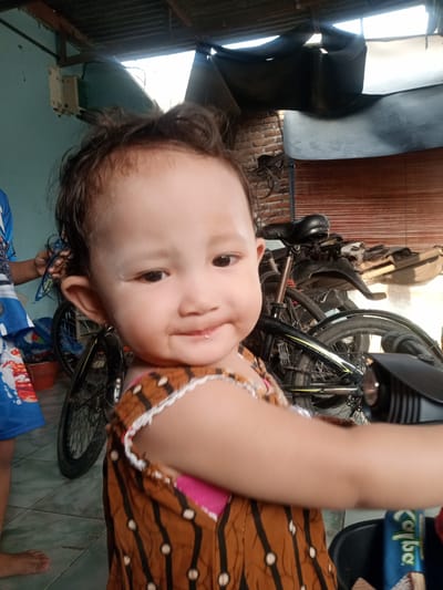 The image depicts a young child with brown hair and eyes, wearing a brown and pink patterned dress with white trim. The child is the primary focus of the image, positioned in the foreground and appears to be smiling. A bicycle is visible in the background, as well as part of another person's blue shirt. A cardboard package is placed on the bottom right. The location appears to be indoors. The building's structure is constructed of exposed brick, wood, corrugated sheets, and some plastered walls. Based on the prompt, the location is Bungur, Indonesia.