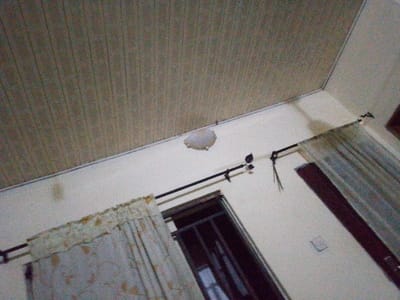 The image shows a low-angle interior view of a room. A paneled ceiling is visible above a wall, which has a window with drawn curtains. A curtain rod is mounted on the wall. A circular indentation is apparent in the ceiling near where it meets the wall. There is discoloration on the wall in several spots.