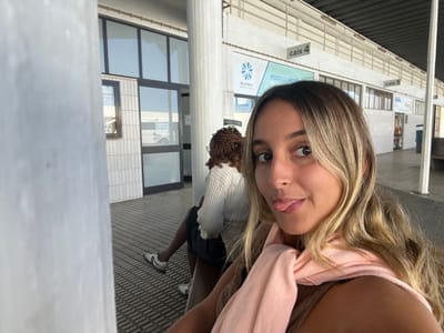 The image depicts a bus terminal in Albergaria-a-Velha, Portugal, under a covered structure with a visible metal roof. In the foreground, a female with long, wavy blonde hair and light skin is seen from the chest up, directly facing the camera with her tongue slightly extended. She wears a light-colored top and a light pink scarf draped around her neck. Behind her and to the left, another female is seated on a bench, viewed from the back and left profile. She possesses dark, possibly braided hair, and is dressed in a white ribbed long-sleeved shirt, dark shorts, and white and black athletic shoes. This individual faces away from the camera, looking towards a building entrance. The building features large glass windows and doors framed in dark material, with the lower exterior section clad in vertical white tiles. Overhead, signs indicating "cais 4" and "cais 5" are visible, alongside a "BUSWAY" logo and partial text "POSTO DE AT..." (likely "Posto de Atendimento"). The ground is paved with patterned grey tiles, and white support pillars are present, one prominently in the left foreground.
