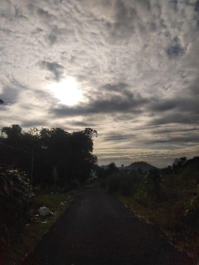 The image displays a dark, asphalted rural road leading into the distance under an overcast sky in Røros, Norway. The sun, a bright, intense white light source, is partially obscured by dense, light-grey stratocumulus clouds in the upper-left quadrant, backlighting the entire scene. The sky, covering approximately two-thirds of the frame, transitions from bright white to various shades of grey, with darker, flatter cloud layers lower down.
The unpaved road, lacking discernible markings, occupies the lower third of the image. Along its immediate left side, a steep, dark embankment is covered in dense vegetation, with scattered patches of light-colored debris visible at its base. Further left, silhouetted by the distant light, are the forms of at least two streetlights amidst dark foliage. The right side of the road is bordered by overgrown, dense green and brown shrubbery and trees. In the background, several dark green hills or low mountains are partially obscured by light mist or low-lying clouds, creating a hazy horizon. No individuals, animals, or vehicles are present. The scene is characterized by a high-contrast lighting condition, with the foreground appearing significantly darker due to the backlight.