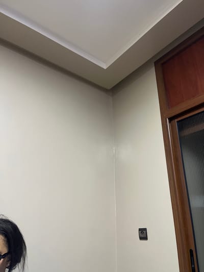 A cream-colored wall meets a white ceiling at a corner. A brown framed window is visible at the top. The wall is plain with a single electrical outlet box. A bit of a person's hair and glasses are visible at the bottom. The lighting casts shadows and highlights across the wall, creating gradients in the cream color.