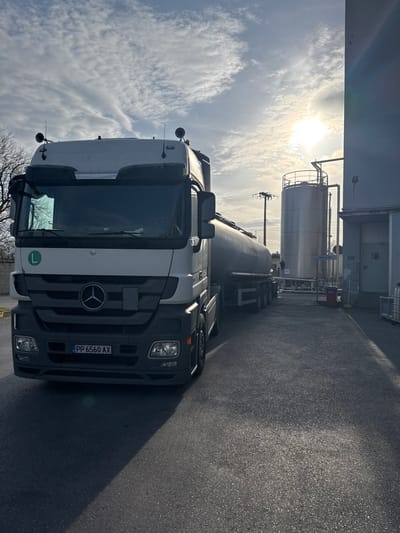 A white Mercedes-Benz Actros tractor unit, bearing a Ukrainian license plate "PP 6560 AX" and a green 'L' learner sticker on its windshield, is parked on an asphalt surface, facing slightly towards the viewer and to the right. It is hitched to a grey, multi-axle tanker trailer, which extends towards the right background. The scene is set in an industrial area within the Municipal Unit of Echedoros, Greece, under a partly cloudy sky with the sun visible and producing lens flare in the upper right. To the right of the truck and trailer, a light-colored industrial building with multiple windows and doors is visible. Adjacent to the building are large, silver cylindrical storage tanks with associated piping and structures. Utility poles with overhead lines are present in the mid-ground. Bare tree branches are visible on the left side, indicating a cooler season. No individuals are present, and no active interactions or specific actions are depicted.