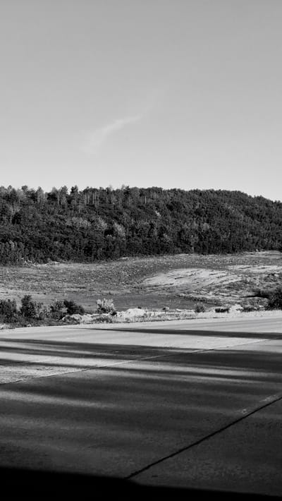 The image is a landscape photograph taken in black and white, oriented vertically. The scene depicts a steep, tree-covered mountainside to the left, with a road running parallel to it on the right. The sky occupies the upper left portion of the image, appearing bright and possibly overcast. The mountainside is densely forested, with numerous trees of varying shades of gray. The ground between the trees is a mix of lighter and darker patches, suggesting rough terrain or vegetation.

The road on the right is paved and appears to be a two-lane highway. Distinct shadows are cast across the road, indicating the presence of a strong light source, likely the sun, though its position is not directly visible. The shadows create a striped pattern on the road's surface. The road appears to be bordered by a rough, rocky embankment on the left side, where it meets the mountainside. A dark, shadowed area is visible in the lower right corner of the image, possibly a structure or a deep shadow cast by an unseen object.

There are no people or discernible objects that would indicate a specific activity or event. The time of day is suggested by the prominent shadows, possibly mid-morning or afternoon. The weather appears to be clear enough for shadows to be cast, but the brightness of the sky might suggest it's not a perfectly sunny day, or that the sky is simply very bright. No text is visible in the image. The overall impression is one of a natural, rugged landscape with a road cutting through it.
