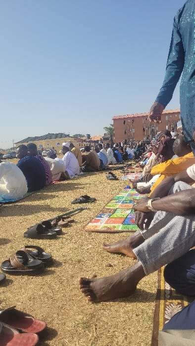 The scene depicts an outdoor gathering of approximately fifty to sixty individuals, predominantly adult males, seated on dry, sparse grass and prayer mats. The event occurs under a clear blue sky, indicating daylight. Most individuals are facing away from the camera, their backs visible, suggesting a collective activity such as prayer. They are dressed in a mix of traditional long garments and casual trousers and shirts. Footwear, primarily sandals, is arranged on the ground in front of many seated individuals; several individuals are barefoot. In the middle ground, on the right side, a multi-colored prayer mat is visible. In the background, several multi-story buildings with light-colored walls and reddish roofs are observed, along with distant vehicles and a slight rise in terrain. The location is Badiko, Nigeria.
