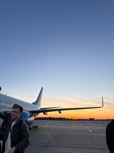 The image shows a man with glasses standing next to a passenger airplane. The airplane has a blue tail fin and a white body. The background shows an airport tarmac and a sunset. The photograph may have been taken at Kitsjanovo airport in Russia.