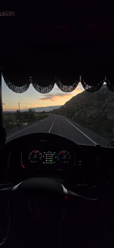 A vertical, eye-level, full shot shows the interior of a truck's cab at dusk. The view is from the driver's seat, looking forward through the windshield. The dashboard is dark and illuminated by various displays. The steering wheel is visible in the foreground. The windshield shows a road winding through a mountainous landscape under a sky with soft orange and pink hues of sunset. A black, fringed fabric hangs from the top of the windshield. The scene appears to be on a road in Bardallur, Spain, given the context provided. The truck's odometer shows 733,098 km. The time displayed is 1:35. The truck is traveling at 75 km/h.
