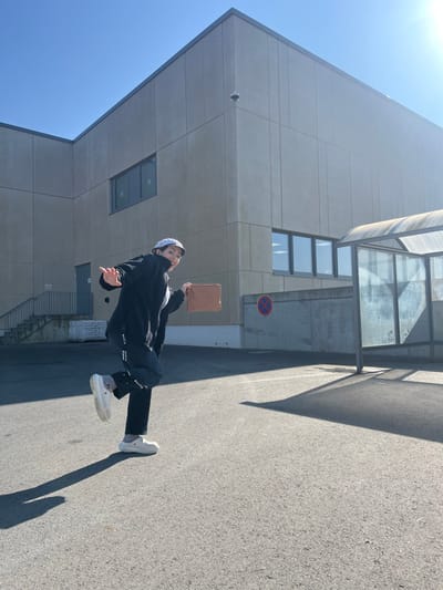 An individual, appearing female, is captured in motion in an outdoor paved area in Roodt-sur-Syre, Luxembourg, under a clear blue sky. The subject wears a light-colored bucket hat, a dark long-sleeved jacket, dark athletic pants with white markings, and white slip-on shoes. She is positioned with her left leg raised and her right arm extended to her left, while holding a light brown rectangular object in her left hand. Her facial expression shows an open mouth. In the background, a multi-story building constructed of light grey concrete panels with dark-framed windows is visible, featuring an external staircase on its left side and a surveillance camera on its upper corner. To the right of the subject, a transparent shelter with a curved roof stands. A red circular "no stopping" sign is affixed to a concrete wall below the building facade. Strong shadows are cast on the light grey paved ground from the subject and the shelter, indicating bright sunlight.
