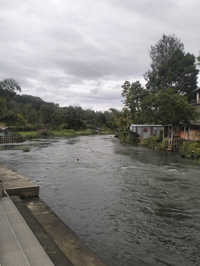 This media file depicts a wide, dark river flowing through a lush, green landscape under an overcast sky. The water is choppy, indicating a moderate current, with ripples and small waves visible. On the left bank, a concrete embankment with steps leads down to the water's edge. A small, weathered wooden structure on stilts is situated near the bank, partially submerged in the water. Further along the river, dense vegetation covers the hills that rise behind it.
On the right bank, several simple structures are built close to the river. One appears to be a corrugated metal building, possibly a dwelling or workshop, with an attached wooden porch or platform. Another structure with an open front and wooden supports is visible next to it. These buildings are surrounded by dense greenery, with tall trees forming a backdrop. The overall setting suggests a rural or semi-rural location, likely in a tropical or subtropical region given the abundant vegetation.
The sky is a uniform grey, suggesting cloudy weather, possibly a dull day or an approaching storm. The light is diffused, casting a soft, even illumination over the scene. There are no people visible in the image, and the focus is on the natural landscape and the man-made structures along the river. The scene evokes a sense of tranquility, though the choppy water hints at underlying energy. The time of day is difficult to determine precisely due to the overcast conditions, but it appears to be daytime. There is no visible text in the image. The location context provided is Blang Bebangka, Indonesia, which aligns with the tropical vegetation and architectural style of the buildings.