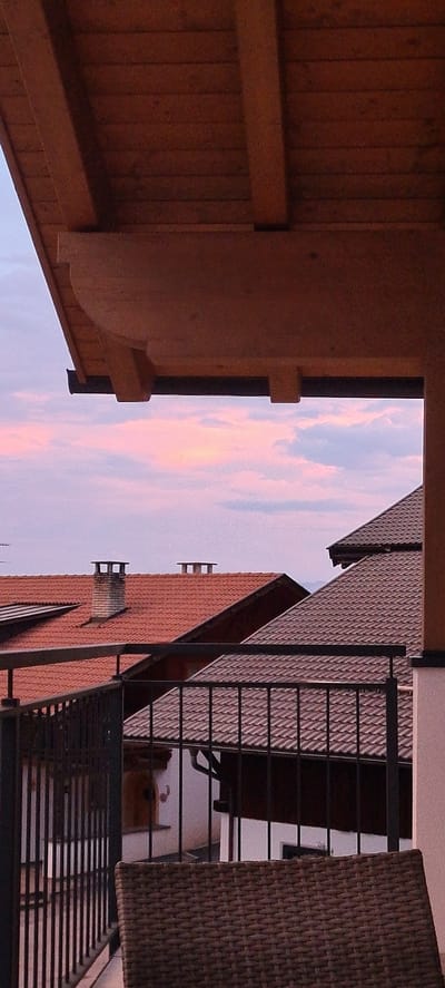 This is a vertically oriented, low-angle, eye-level outdoor photograph taken during dusk. The scene looks out from a wooden covered porch towards a residential building with red tiled roofs and a balcony with a black metal railing. The sky is filled with soft pink and purple clouds, indicative of sunset or sunrise.

Prominently featured in the foreground on the left is a section of the wooden porch ceiling, constructed with parallel wooden planks and supported by thick wooden beams. To the right of the porch structure, the residential building's architecture is visible, including a steeply pitched red tile roof with two rectangular ventilation elements protruding from it. A balcony with a black metal railing overlooks the scene. Part of a woven outdoor chair or seating element is visible on the far right edge of the frame.

The time of day is clearly indicated by the colorful sky. The weather appears clear, with the clouds being the only atmospheric element. The architectural style of the building, with its red tiled roof, suggests a European location. The presence of the porch and balcony implies a residential setting, likely a home or a small apartment building. No people are visible in the image. No text is present. The overall mood is calm and serene, likely due to the soft lighting of dusk and the peaceful view.