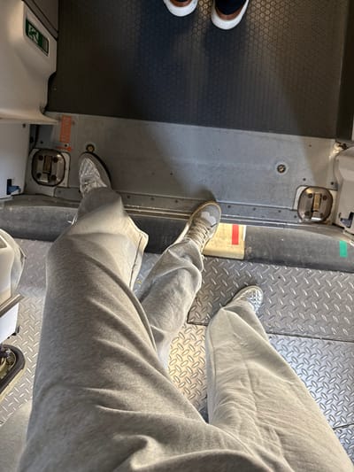 The image depicts an interior view from a downward angle, showcasing a person's lower body and feet, along with the feet of two other individuals, within what appears to be an aircraft cabin. In the foreground, a person's legs are clad in grey sweatpants, their feet resting on a grey metal diamond plate floor. The left foot wears a light-colored sneaker with a quilted texture, while the right foot wears a light tan and grey sneaker.

Directly beyond the foreground feet, the floor transitions to a flat grey metal surface featuring floor latches or mounts and an orange "ARMED" label with an arrow pointing to a mechanism on the left. A white panel with a green exit pictogram is visible to the upper-left. Further back, two additional pairs of feet are visible, standing on a dark grey rubber floor mat with a repeating hexagonal pattern. These individuals wear dark shoes with light-colored soles.

The presence of aircraft-specific door mechanisms, exit pictograms, and multi-textured flooring indicates an airplane interior. This scene is located at the Tijuana International Airport in Tijuana, Mexico.