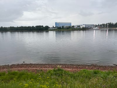 The image shows a wide river flowing in front of an industrial area with buildings and a large shipyard. The sky is overcast with grey clouds. The riverbank in the foreground is covered with green grass and a rocky, reddish-brown shore. In the distance, a line of trees separates the river from the industrial complex. A prominent blue and white building with a tall chimney stands out. Several ships, including a white yacht and a tall-masted vessel, are docked at the shipyard. Reflections of the buildings and lights shimmer on the water's surface. The scene appears to be a view of the Neva River near the town of Imeni Sverdlova, Russia, a location known for its industrial and shipbuilding activities.