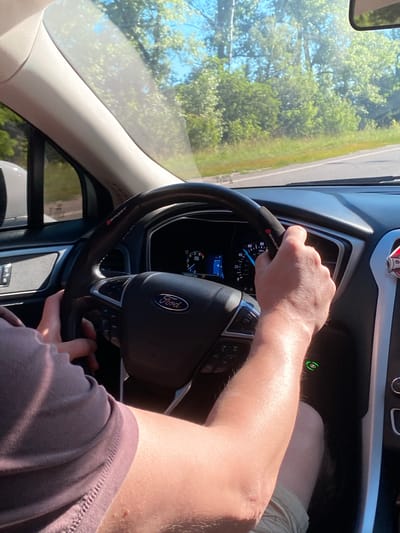 The image shows the interior of a Ford vehicle driven by a person. The driver's hands are visible on the steering wheel, which has a "SPORT" marking. The dashboard displays a speedometer with a reading around 40-50. Outside the car, a sunny day with green trees and a road can be seen through the windshield. The location is stated to be Herasymivka, Ukraine, but there is no visual information in the image to confirm this specific location.