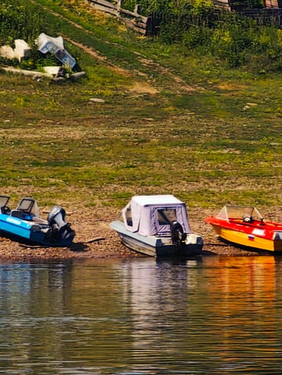 The image depicts a waterside scene in Pogodaevo, Russia, characterized by a calm body of water in the foreground and a grassy, slightly overgrown embankment in the background. Three boats are visible resting on the rocky shore. The leftmost boat is a blue motorboat with a visible outboard motor. Adjacent to it, to the right, is a small boat with a grey canopy. Further to the right, a red and yellow motorboat with a clear windshield is positioned. The embankment behind the boats features sparse vegetation and a wooden fence structure at the top. The water reflects the surrounding environment, creating a textured surface. There are no people visible in the frame.