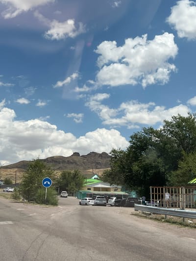 The image depicts a roadside scene in Karlygash, Kazakhstan. The sky is blue with scattered white clouds. In the foreground, a paved road curves to the right. To the left of the road, a round blue traffic sign with white arrows pointing left and right is visible, surrounded by green foliage. Further ahead, several cars are parked or driving on the road, including white sedans and darker SUVs. Beyond the road, a few low-lying buildings with green and light-colored roofs are situated. A line of trees runs along the middle ground, separating the road area from a rocky, mountainous landscape in the background. To the far right of the image, a metal fence encloses an area with some individuals standing near what appears to be a shaded structure.